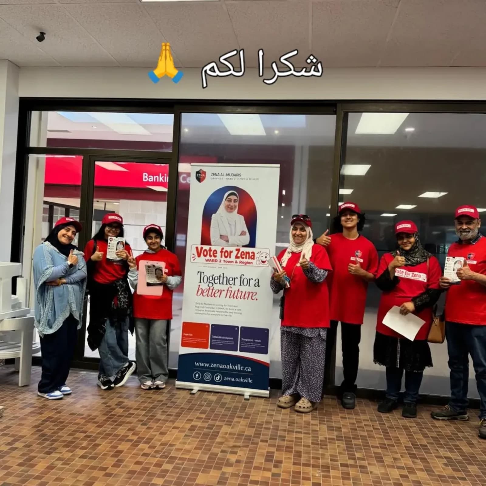 Campaign volunteers in red Vote for Zena shirts and caps giving thumbs up beside the campaign banner in an Oakville plaza